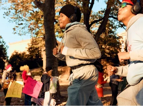Two runners in the foreground with headphones, while spectators with signs are visible in the background.