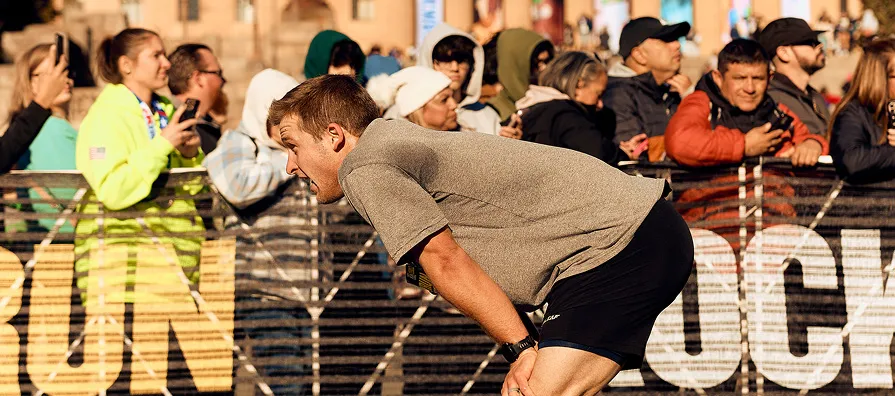A male runner bends over, catching his breath, with spectators watching from behind a barrier.