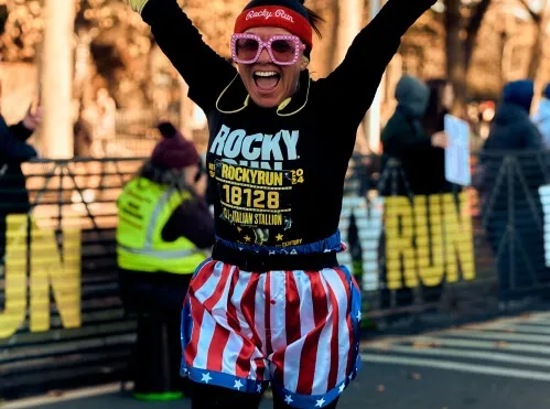 A runner in a Rocky Balboa-inspired costume, complete with American flag shorts and pink sunglasses, celebrates with arms raised at the Rocky Run.
