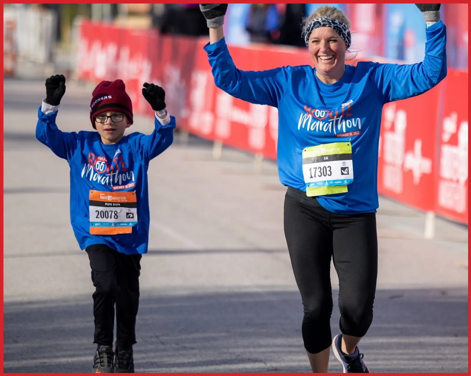 A young boy and an adult woman, both wearing blue race shirts and with their arms raised in celebration, run towards the finish line at the Route 66 Marathon.