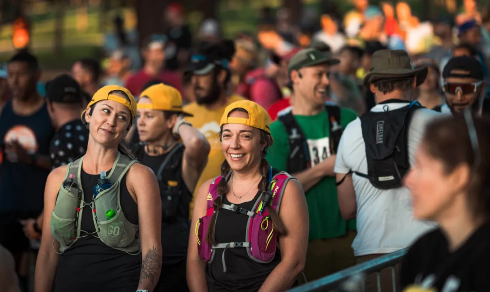 Two women in yellow hats smiling at the start of a race, with a crowd of other runners in the background.