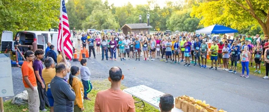 A large group of runners gathered at the start line of a race, with an American flag and tents in the background.