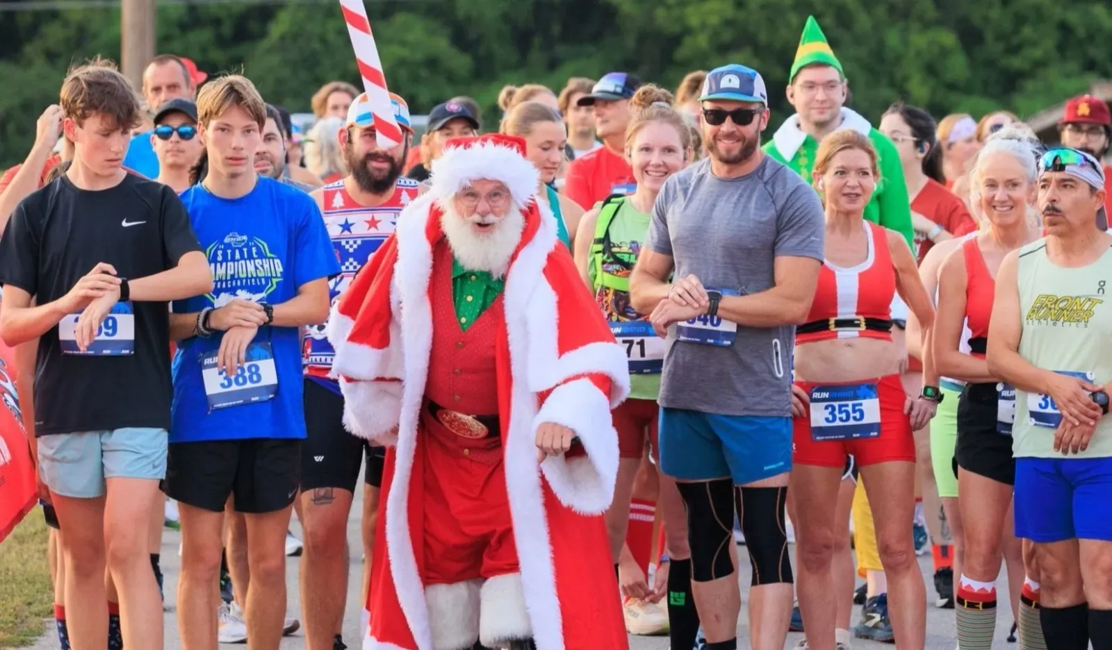 A group of runners, including Santa Claus and an elf, stand at the start line of a race, smiling and checking their watches.