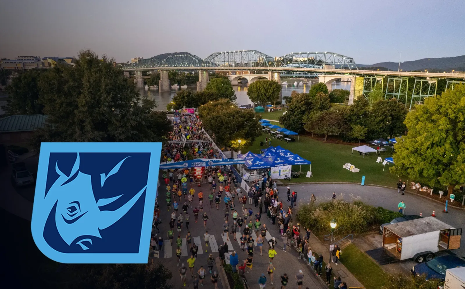 A large group of runners crossing the start line of the 7 Bridges Marathon, with a scenic bridge and river in the background.
