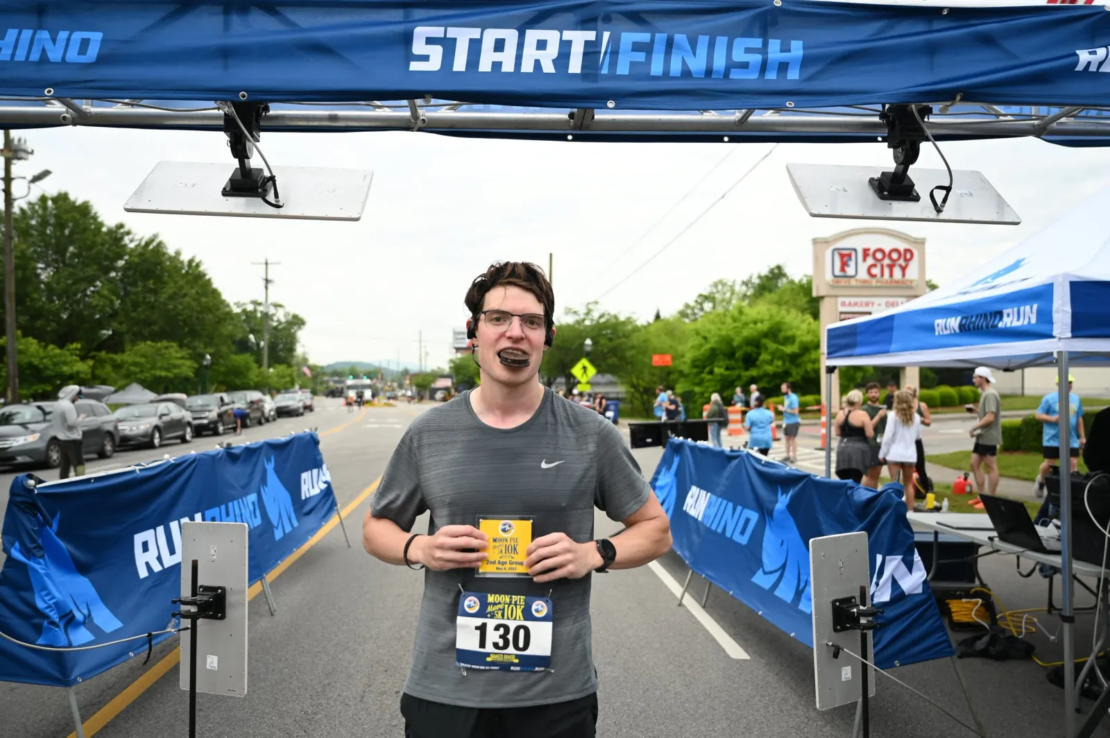 A runner smiles at the camera, holding a bib and a small award, with a 'START/FINISH' banner overhead.