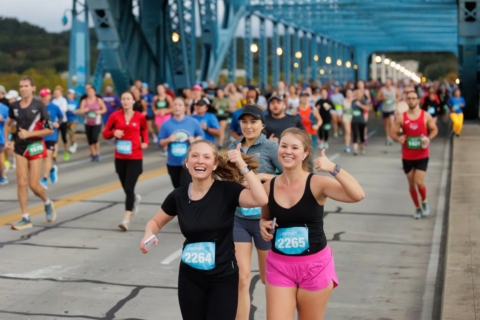 Two smiling female runners giving thumbs up while participating in a race on a bridge, with a large crowd of other runners behind them.