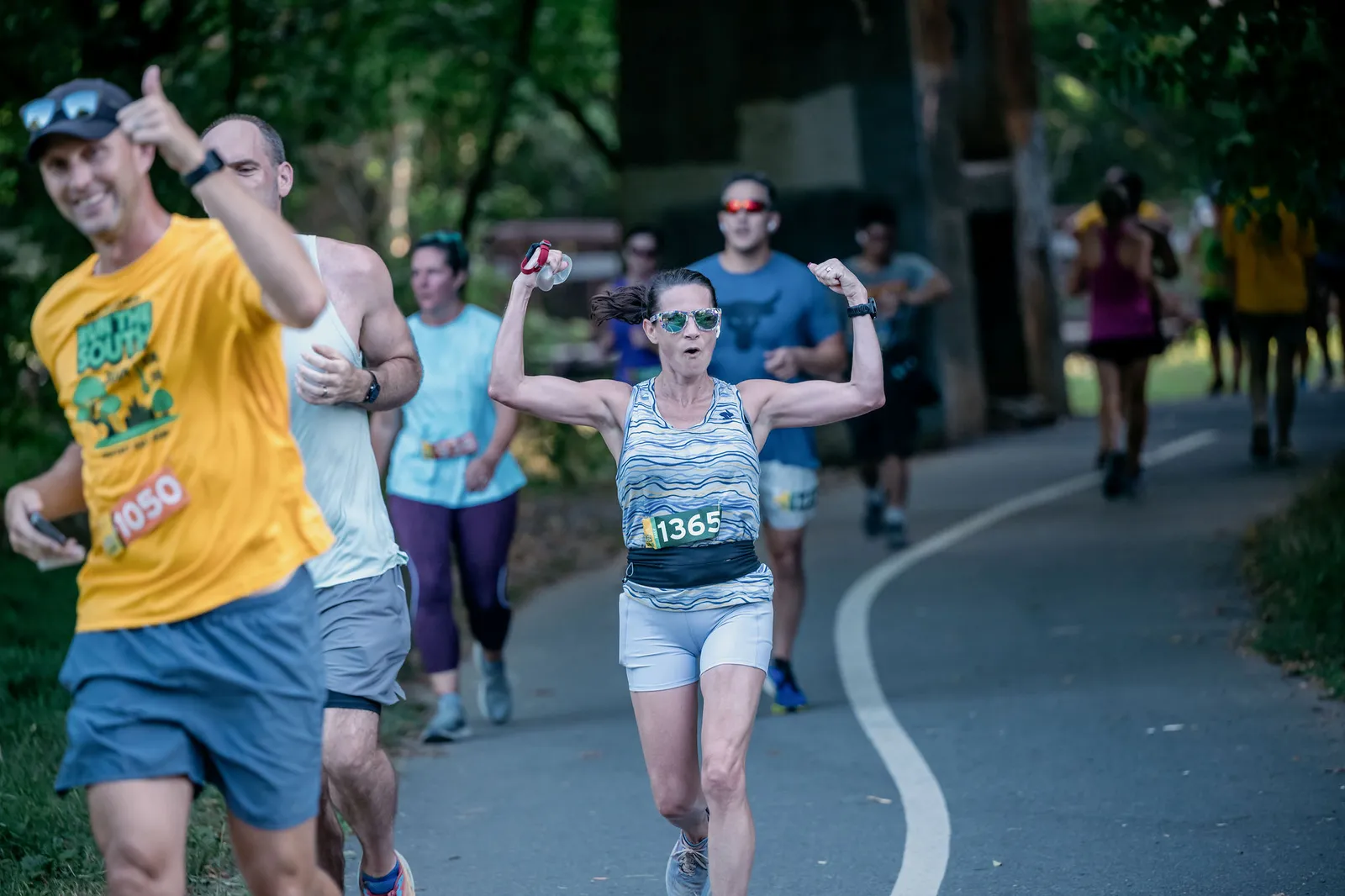 A female runner with bib number 1365 flexes her arms triumphantly while running on a paved path, with other runners visible in the background.