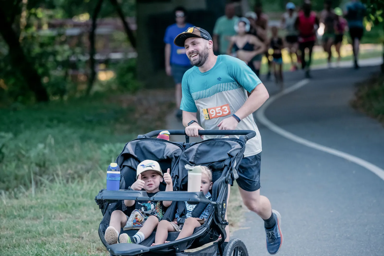 A man runs with a double stroller containing two young children during a race, with other runners in the background.