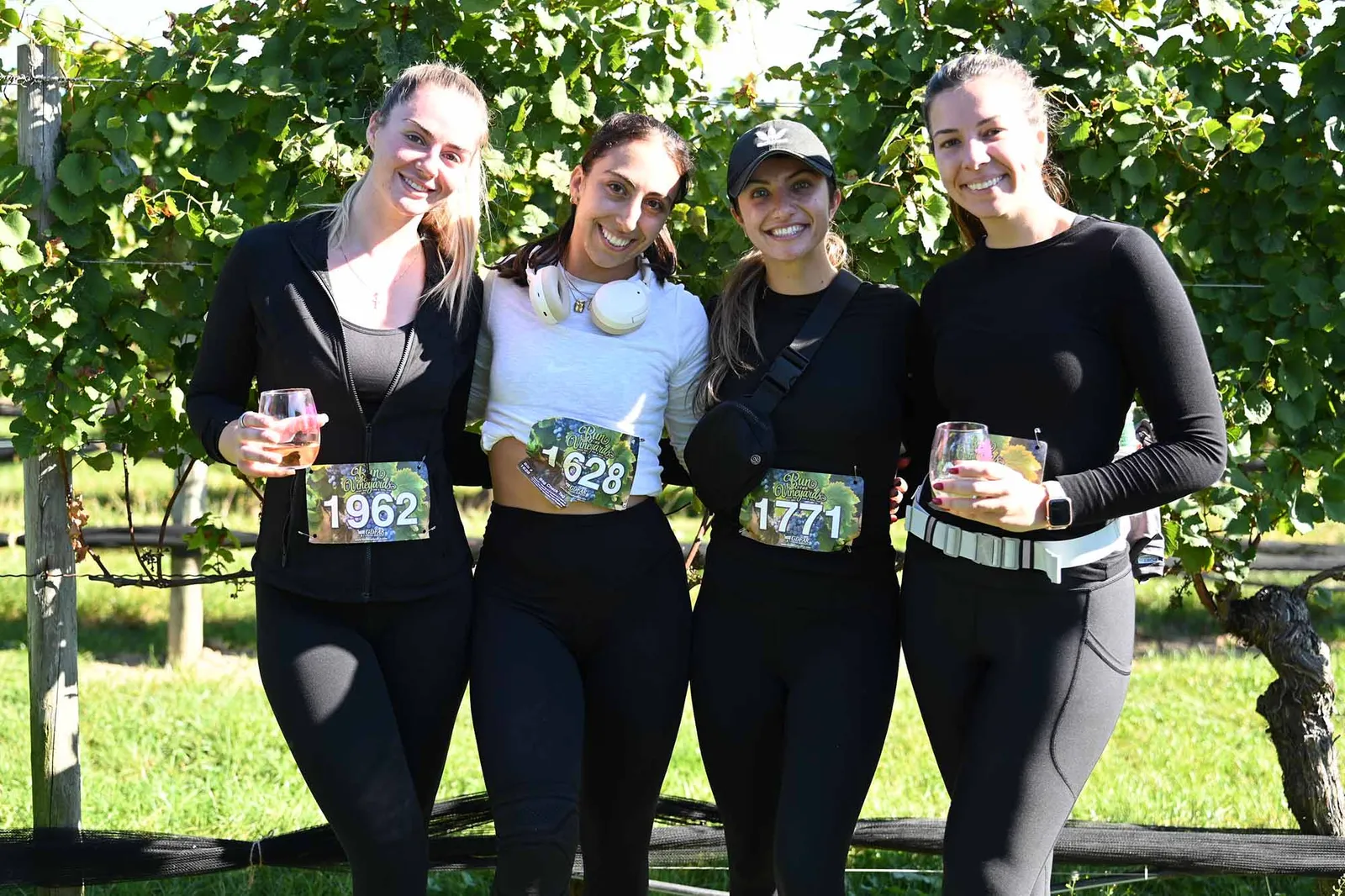 Four smiling women wearing race bibs and holding wine glasses in front of a vineyard.