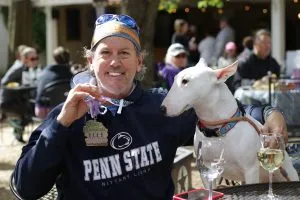 A smiling man wearing a Penn State hoodie and a race medal, posing with a white bull terrier at an outdoor event.