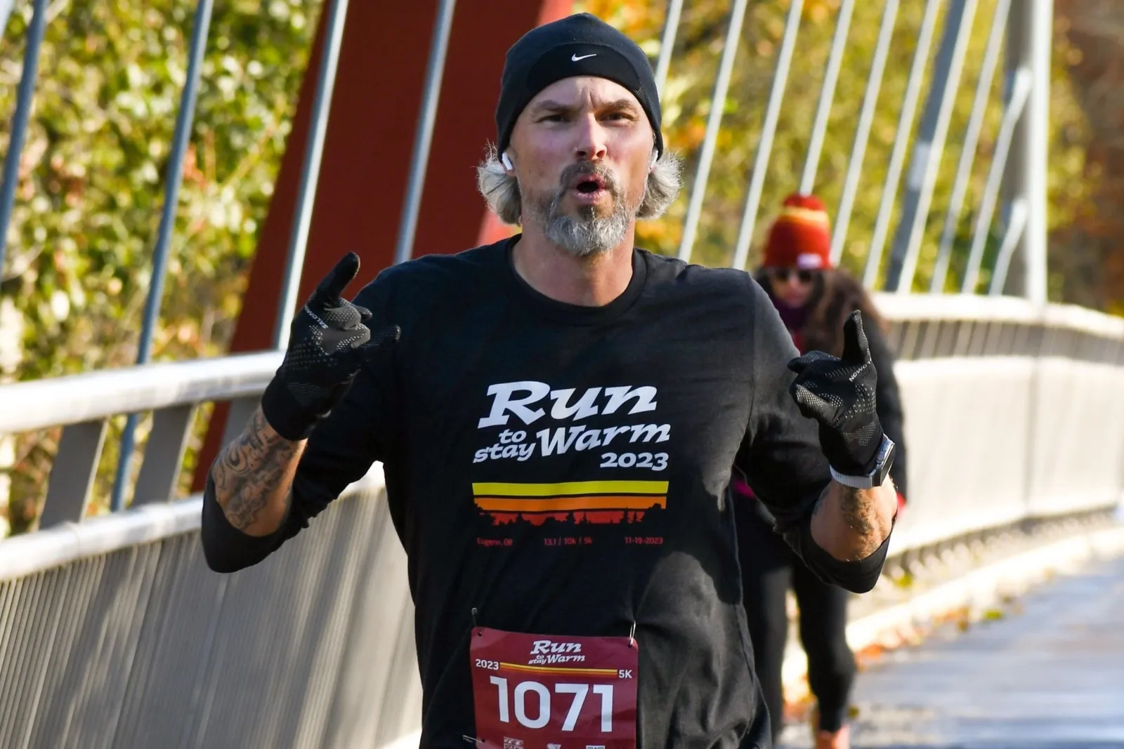 A male runner with a beard and beanie runs across a bridge, wearing a black t-shirt with 'Run to Stay Warm 2023' printed on it and bib number 1071.