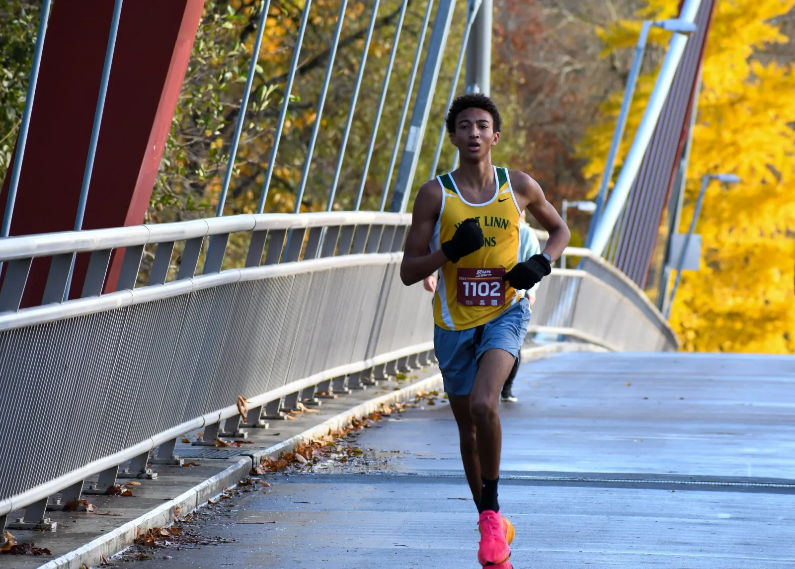 A young male runner in a yellow and green singlet runs across a bridge, with vibrant yellow trees in the background.