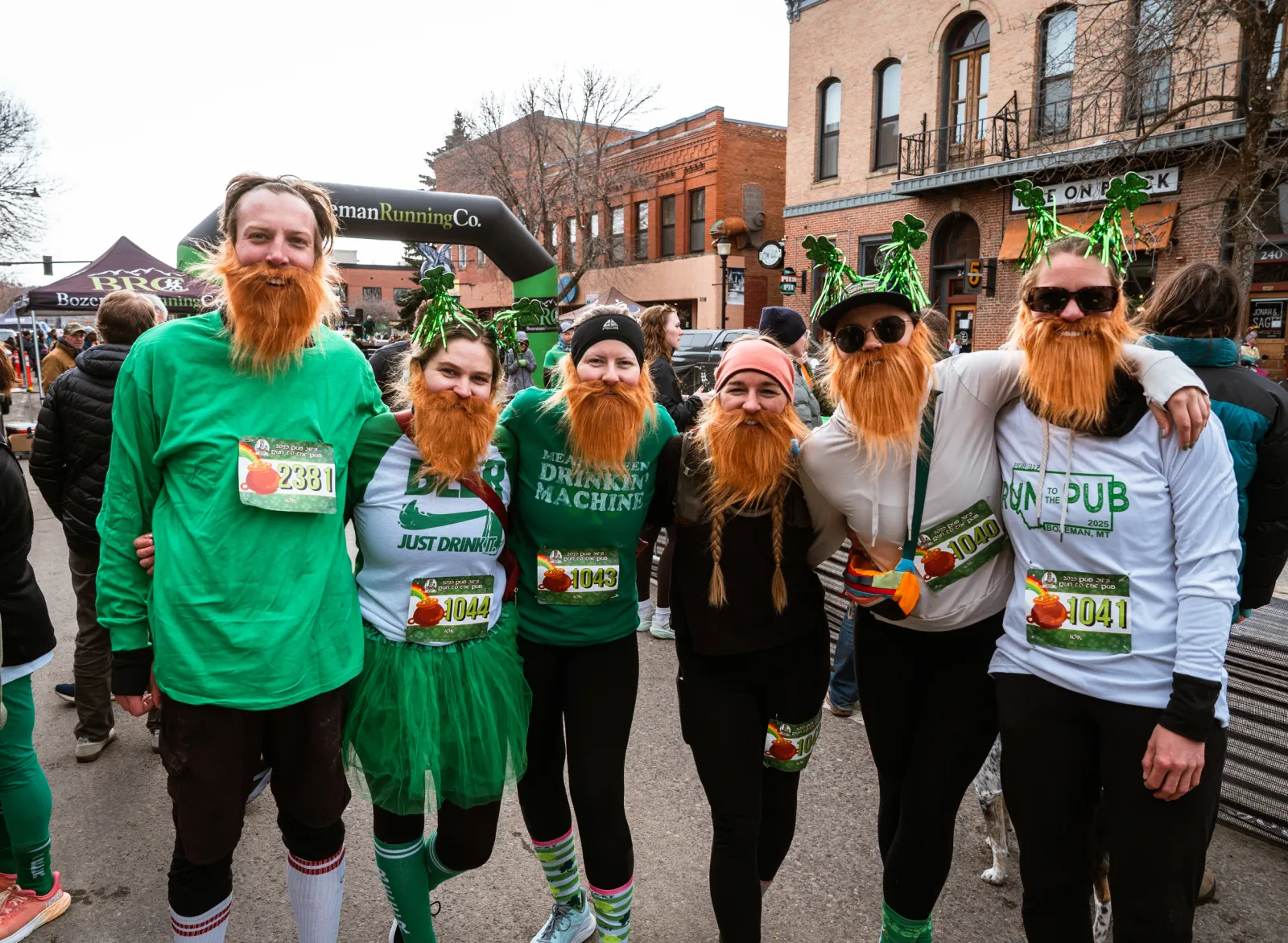 A group of runners wearing fake beards and St. Patrick's Day themed hats smile at the camera in front of a race archway.