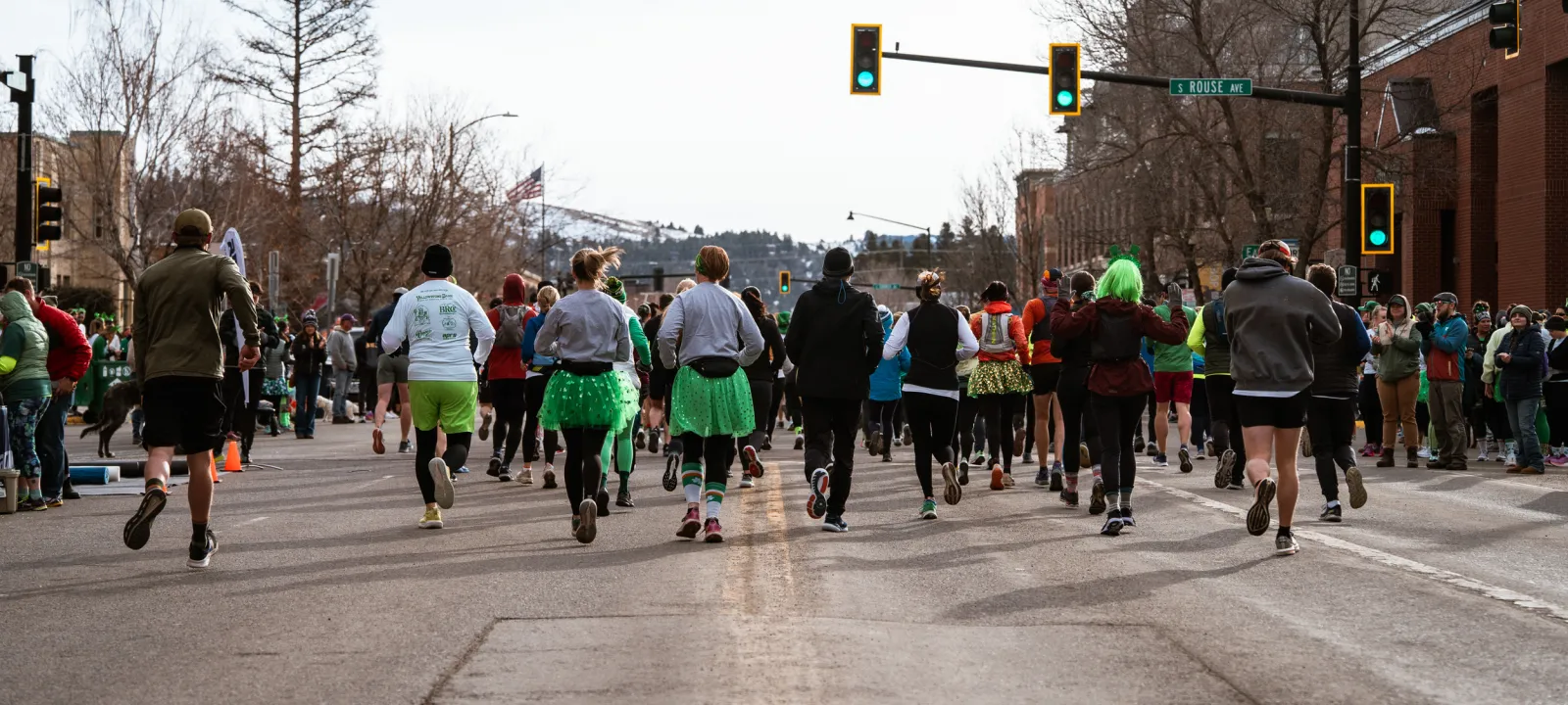 A group of runners, many wearing green tutus and hats, run down a city street during a race.