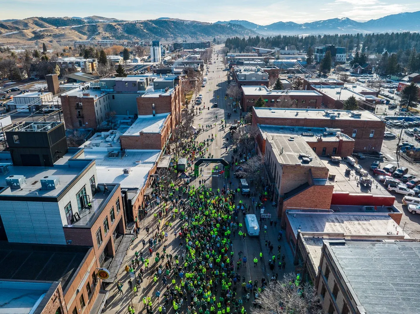 Aerial view of a large crowd of runners gathered on a city street, with an archway at the start line and mountains in the distance.