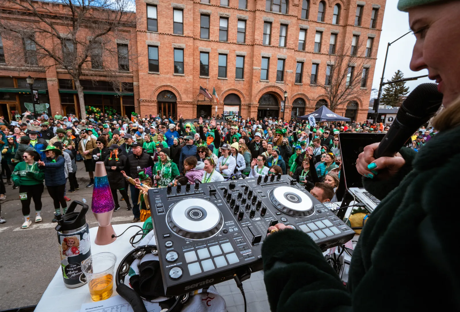 A large crowd of people, many wearing green, gathered in front of a brick building, with a DJ setup in the foreground.