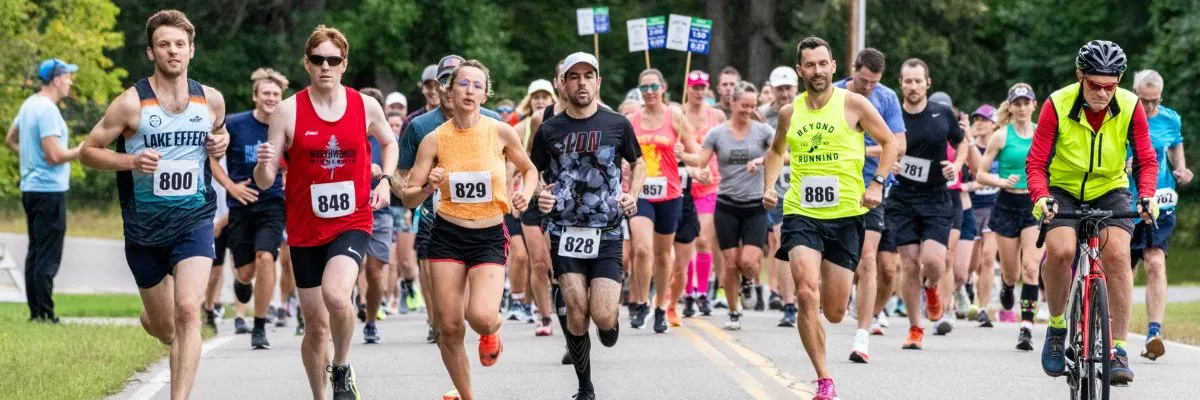 A group of runners of diverse ages and genders are running down a road, with trees and foliage in the background. Some runners have bib numbers visible.