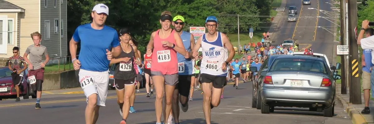Runners participating in a race, with bib numbers visible, on a road with spectators and cars in the background.