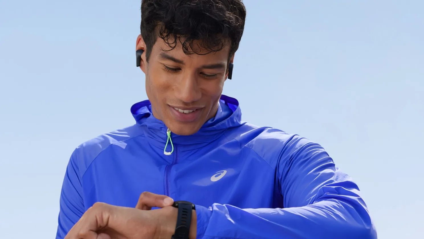 A runner in a blue jacket checks their watch against a clear blue sky.