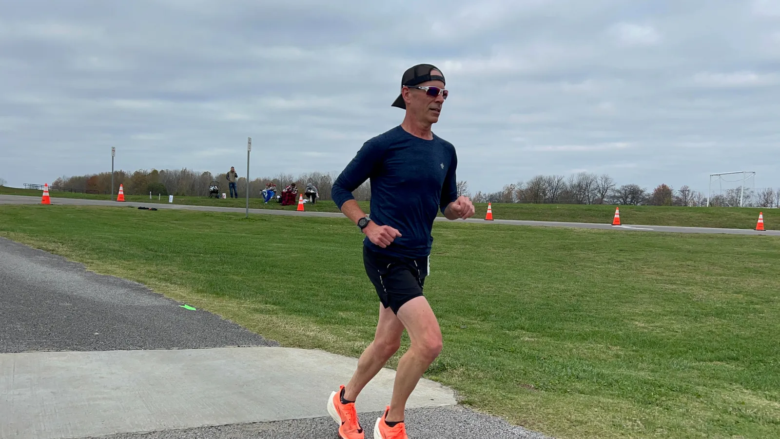 A male runner in a dark blue shirt and black shorts runs on a paved path with a grassy field to his right.