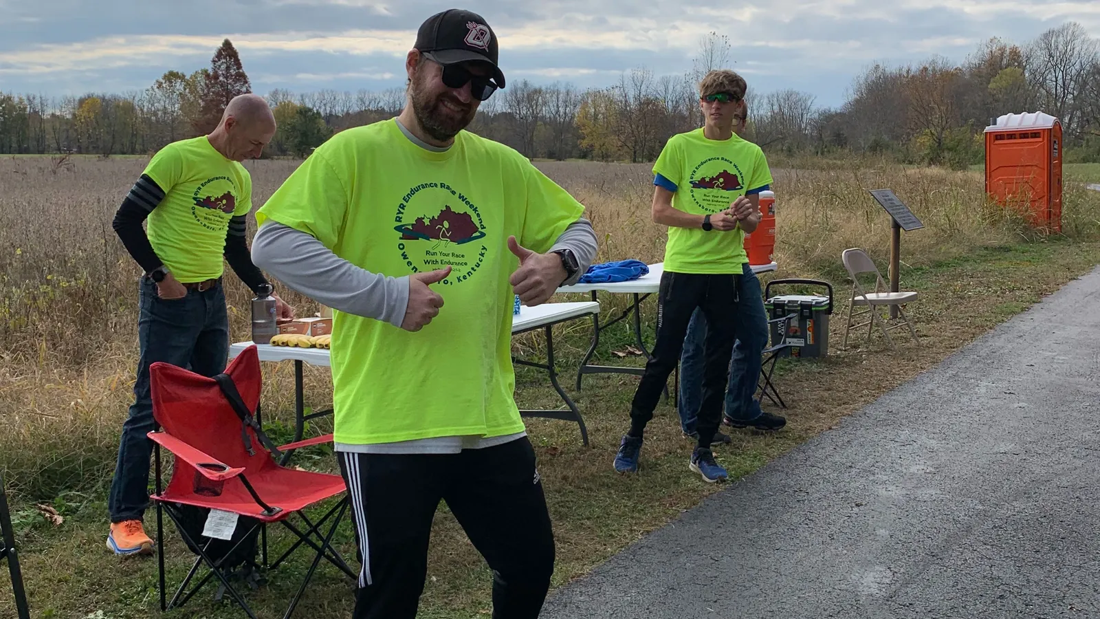 Three people wearing bright yellow race t-shirts stand near tables with drinks and snacks, with one person giving a thumbs up.