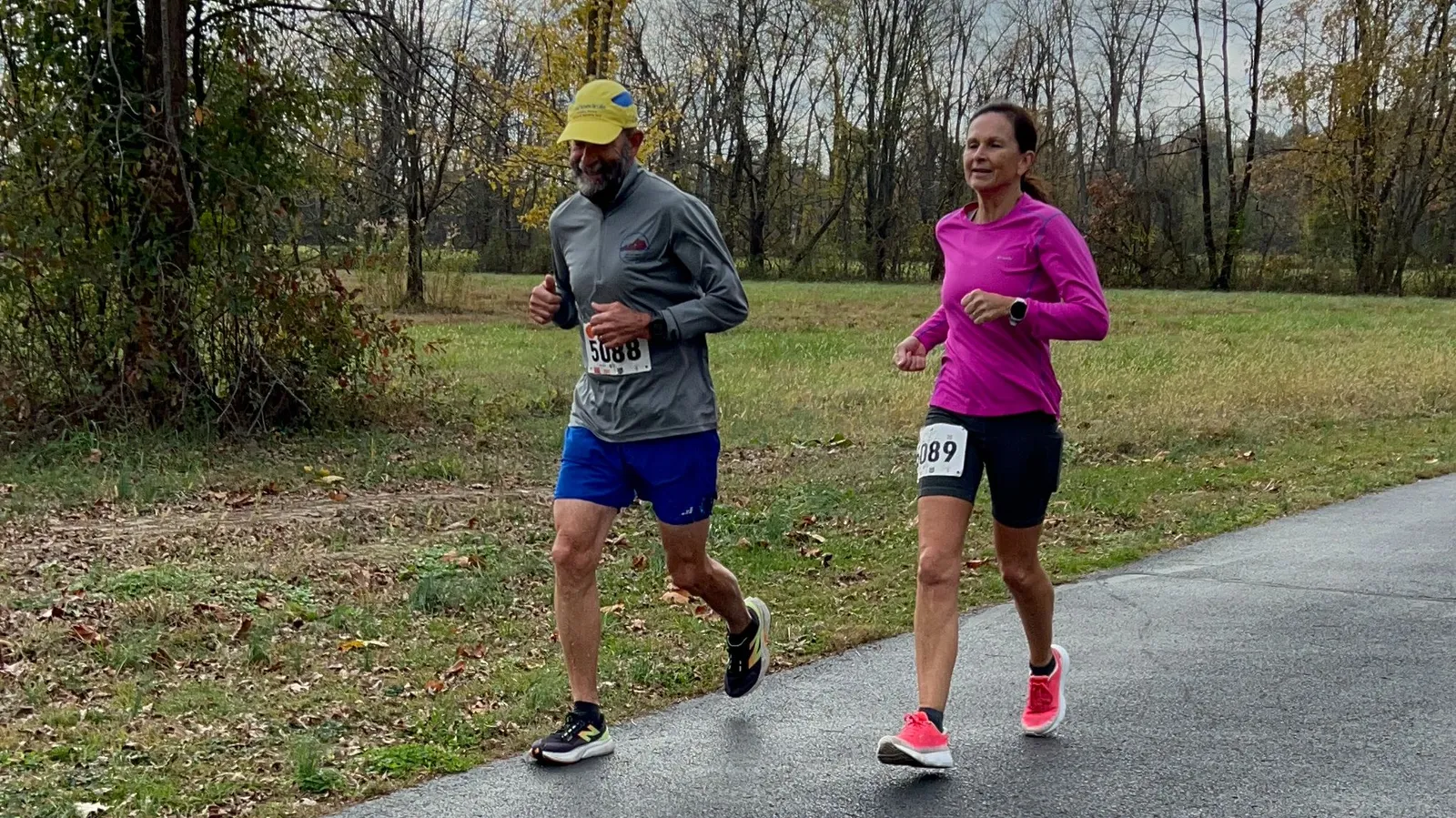 Two runners, a man and a woman, running on a paved path in a park-like setting.