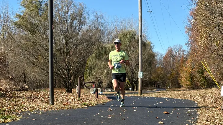 A male runner wearing a light green t-shirt with bib number 4918, black shorts, and a cap runs on a paved path lined with trees and fallen leaves.
