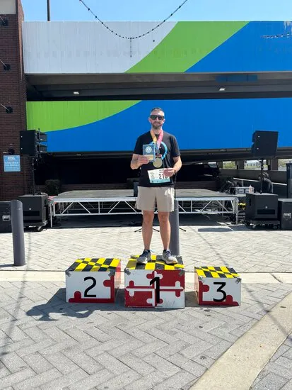 A runner stands on the first-place podium, holding a medal and a certificate, with a backdrop of a large mural and stage.