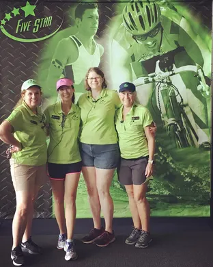 Four women in green shirts and shorts stand together in front of a green and black backdrop featuring images of a runner and a cyclist, with the words 'Five Star' visible.