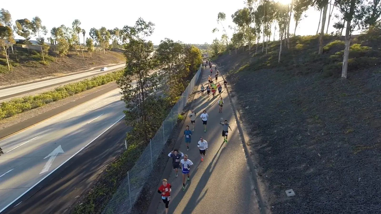 Runners on a paved path next to a highway, with trees and hills in the background.