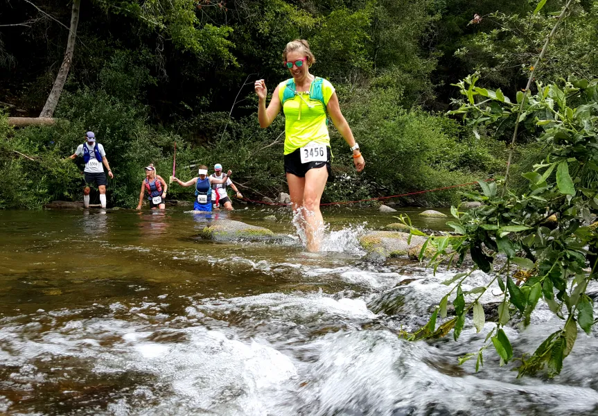 A female runner in a bright yellow shirt and black shorts splashes through a shallow river during a trail race, with other runners visible behind her in the water.