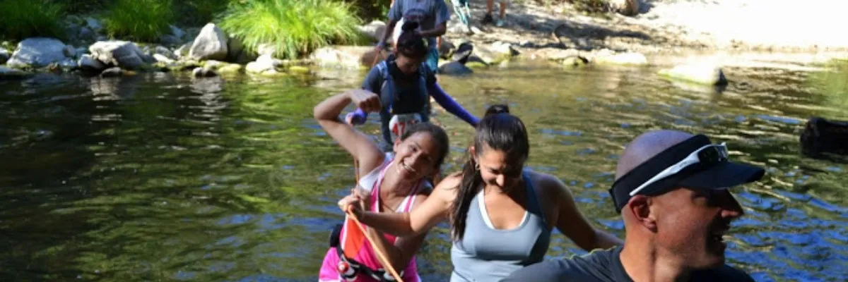 Runners crossing a shallow river on a trail, with one runner in the foreground smiling and holding onto a rope.