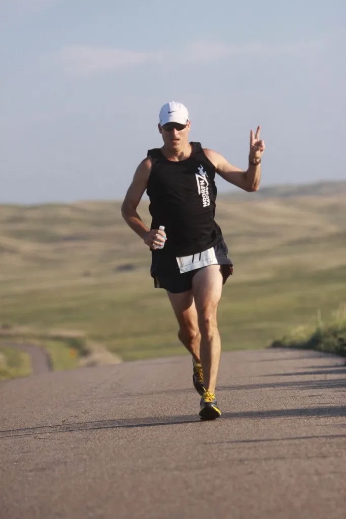 A male runner wearing a black tank top and a white cap runs on a paved road with rolling hills in the background. He is holding a water bottle and giving a peace sign with his right hand.