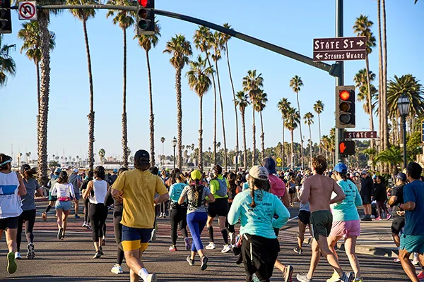 Runners participating in the Santa Barbara Half Marathon on a sunny day, with palm trees lining the road and a street sign indicating State Street and Stearns Wharf.