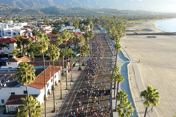 Aerial view of a large marathon race with many runners on a road lined with palm trees, next to a beach and the ocean.
