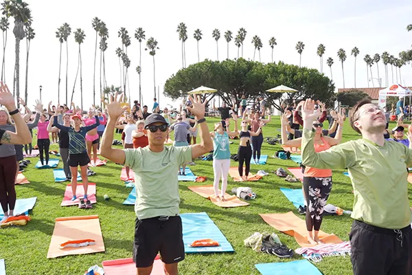 A large group of people practicing yoga on mats on a grassy field with palm trees in the background.