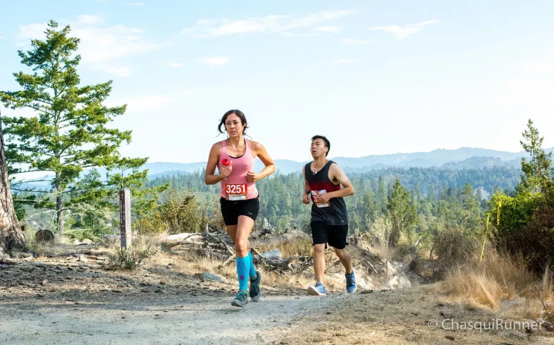 Two runners, a woman in the foreground and a man behind her, running on a dirt trail with a scenic view of forested hills in the background.