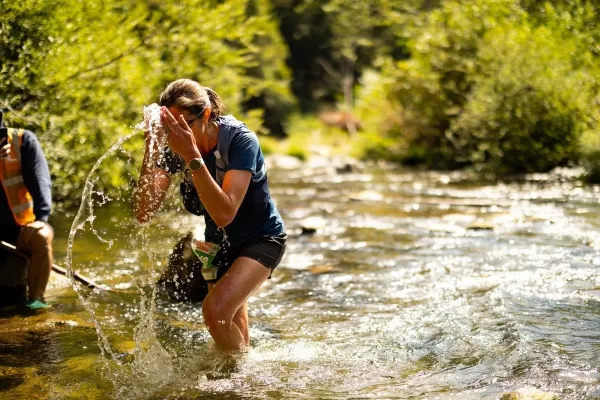 A female runner splashes water on her face while wading through a stream during a trail race.