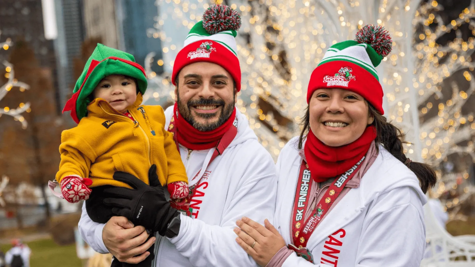 A smiling family, including a baby dressed as an elf, wearing Santa hats and race medals, posing in front of festive holiday lights.