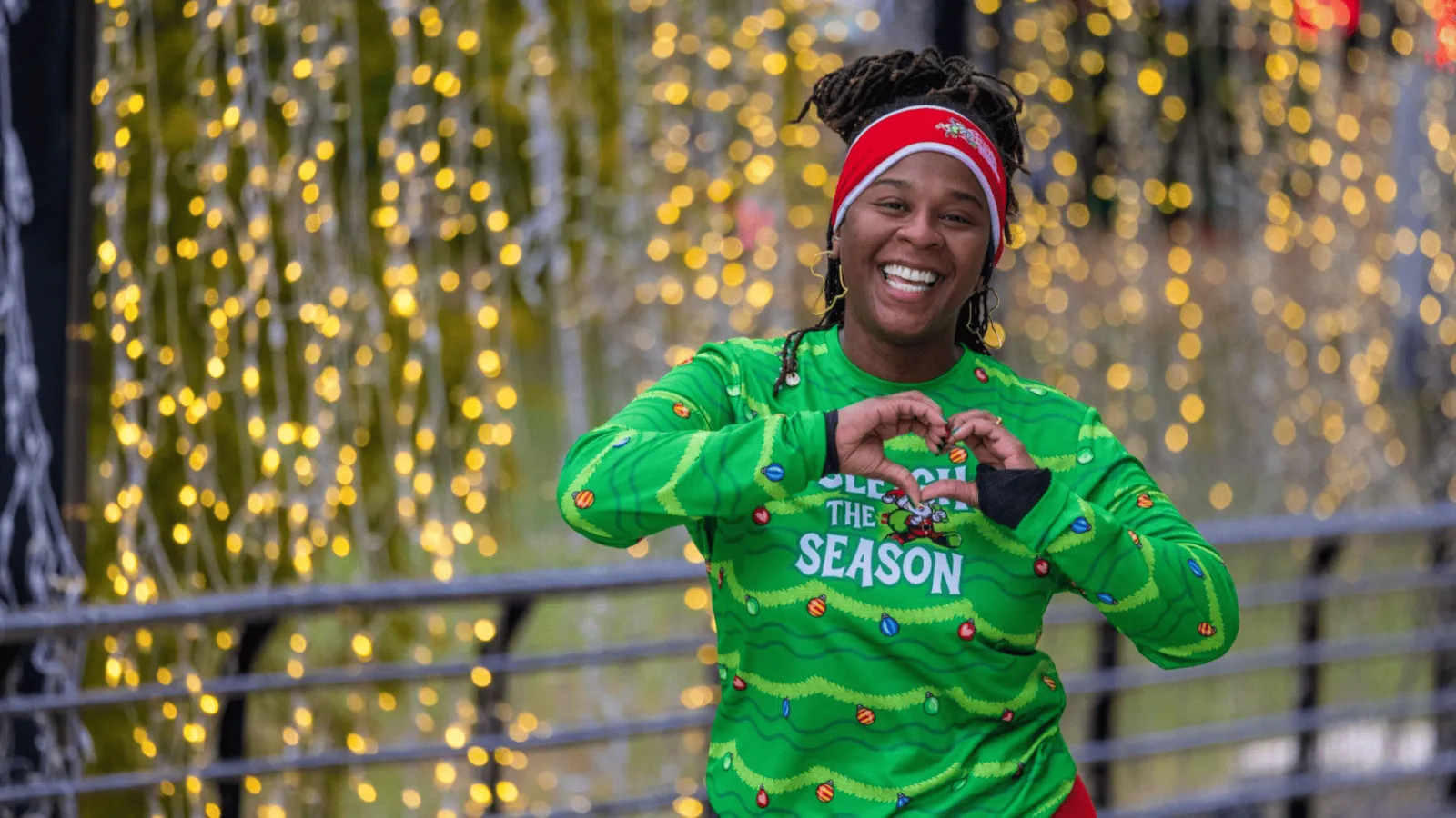 A smiling woman wearing a green long-sleeved shirt with Christmas lights and ornaments, and a red headband, makes a heart shape with her hands in front of a background of blurred yellow lights.