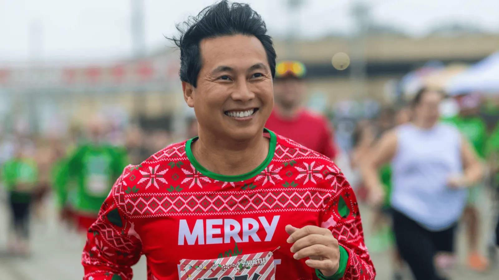 A smiling man wearing a festive red 'Merry' sweater runs in a race with other participants in the background.