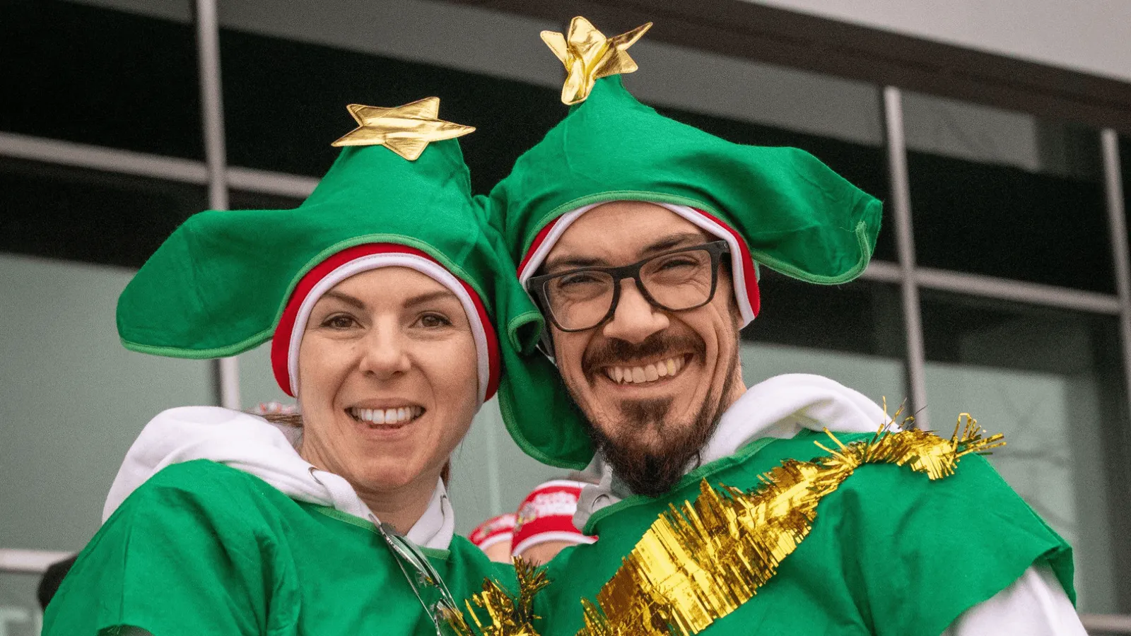 Two smiling people wearing festive green elf hats with gold stars and green elf costumes, celebrating at a race.