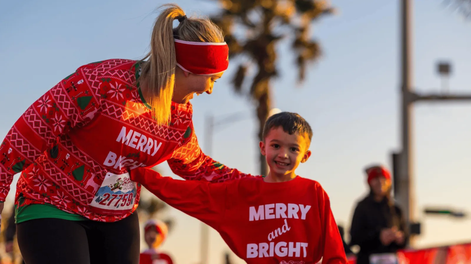 A woman in a festive red sweater helps a smiling young boy in a red shirt during a race, with a palm tree and clear sky in the background.