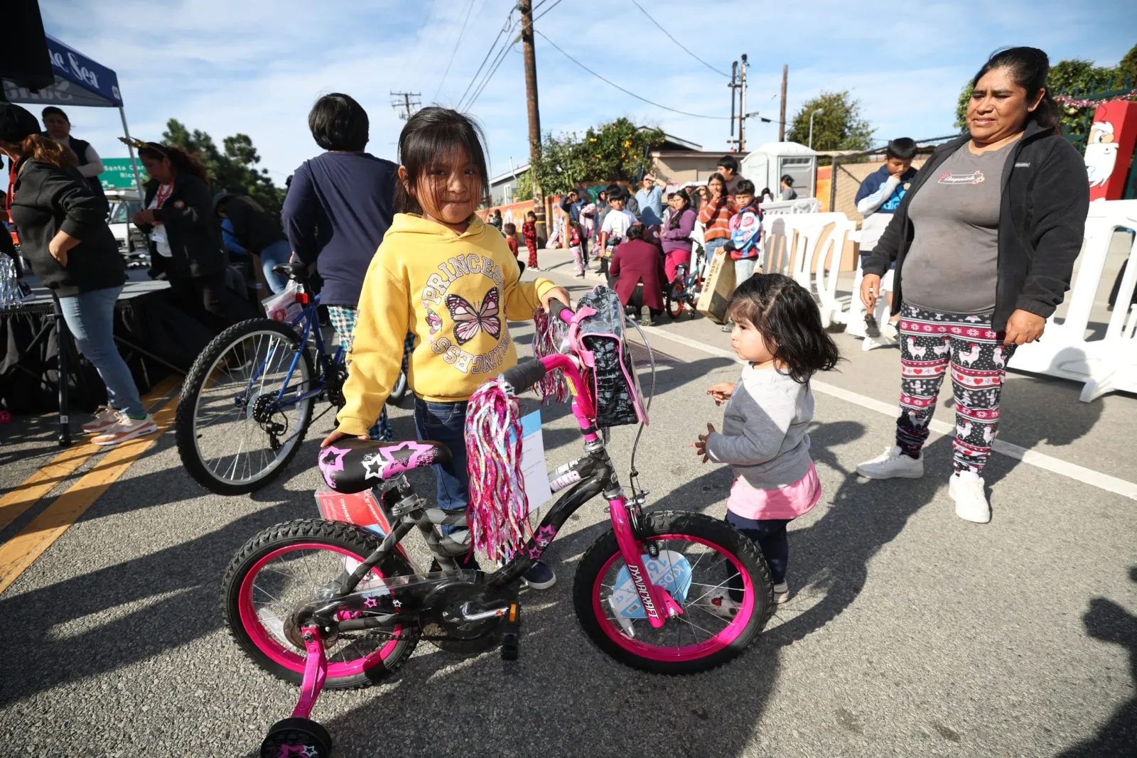 A young girl in a yellow hoodie stands with a pink bicycle, looking towards the camera, while other people and a woman in patterned leggings are visible in the background at an outdoor event.