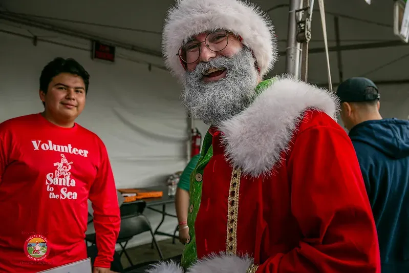 A volunteer in a red 'Santa to the Sea' shirt stands next to a person dressed as Santa Claus.