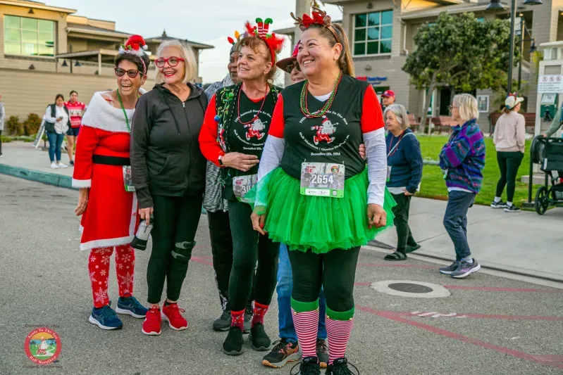A group of women dressed in festive holiday costumes, including Santa outfits and elf tutus, stand together at what appears to be the start of a race, smiling and looking towards the camera.