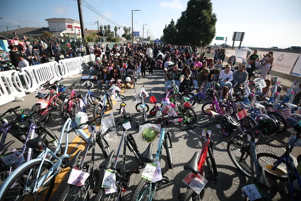 A large crowd of people, including many children, gathered around a street lined with numerous bicycles, suggesting a giveaway or community event.