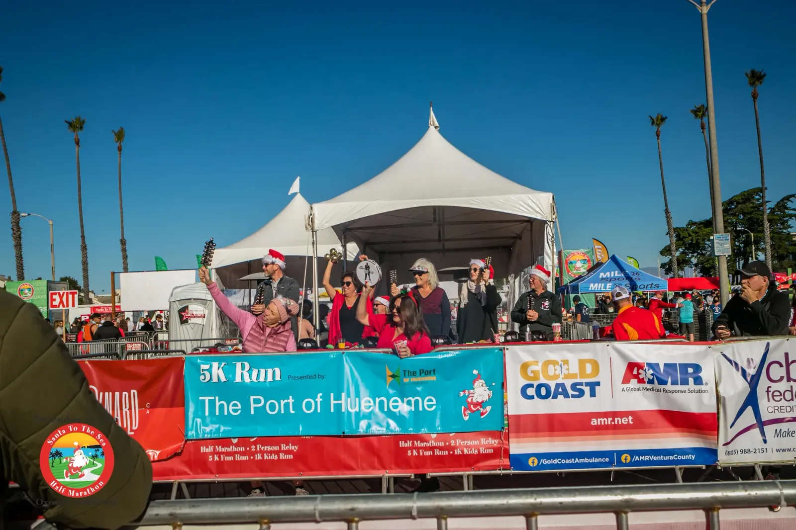 People in Santa hats celebrating at a race event, with banners for '5K Run The Port of Hueneme' and 'Gold Coast AMR'.