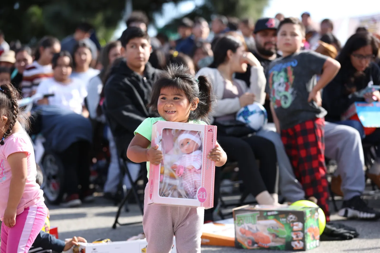 A young girl smiles while holding a doll in a box, with a crowd of people in the background.