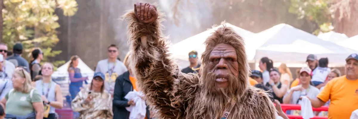 A person in a Bigfoot costume with their fist raised in celebration, surrounded by a crowd at an outdoor event.